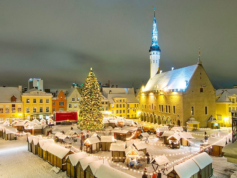 An image of a Christmas market, with tiny stalls covered in snow, a large decorated Christmas tree covered in lights, and a town hall and steeple.